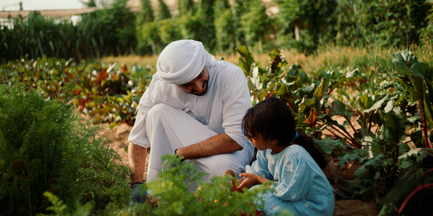 Man in traditional white clothing and a young girl in a blue dress kneeling and gardening together in a lush vegetable garden.